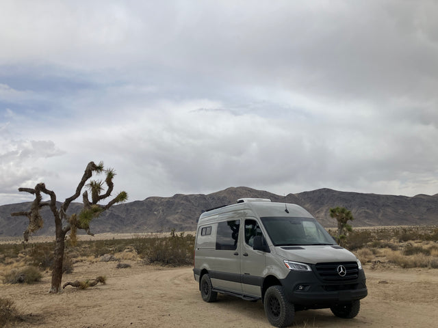Explore the desert! This adventure camper van, ideal for RV rental, is parked near a Joshua tree with mountains under cloudy skies.