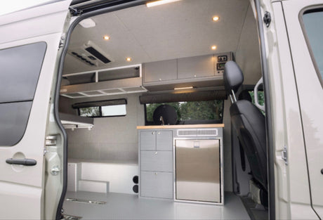 rv rental camper van interior. Features kitchenette with fridge, overhead cabinets, bed, and bright lighting.