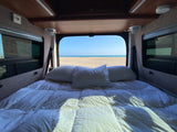 Cozy rv rental bed with white linens and pillows, framed by the open back, reveals a stunning sandy beach & clear ocean view.