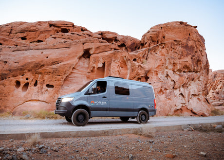 A grey Moterra camper van RV rental on a dirt road, parked by stunning red rock formations in a desert landscape.