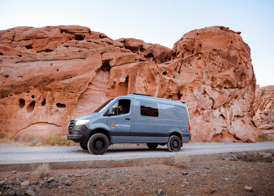 A grey Moterra camper van RV rental on a dirt road, parked by stunning red rock formations in a desert landscape.