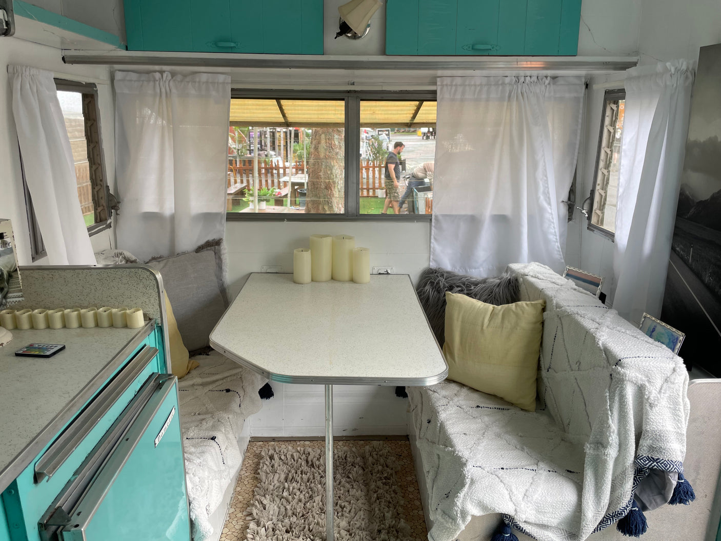 Cozy RV rental interior with a dining area, white curtains, light blue cabinets, and a view of people outside highlight this camper.