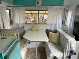 Cozy RV rental interior with a dining area, white curtains, light blue cabinets, and a view of people outside highlight this camper.