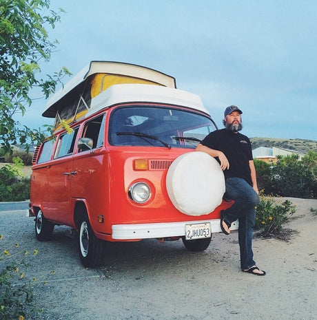Bearded man leaning on a vintage orange VW camper van with a pop-top, ready for an RV rental road trip adventure!