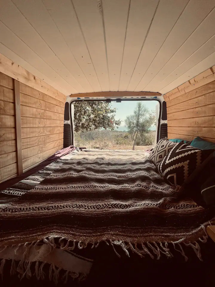 Inside a wooden-paneled camper van. The bed has a patterned blanket, looking out to a scenic view. Ideal RV rental. — photo 8