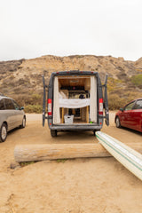 An RV rental camper van, doors open, revealing a cozy bed. Parked on a sandy beach with a surfboard, other cars, and cliffs.