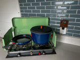 Compact RV rental kitchen: green portable camping stove with a blue pot & black pan on a gray counter.
