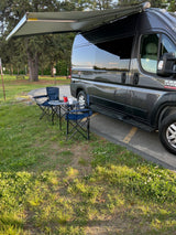 Grey RV rental van with extended awning, two blue chairs, and a table set up on the grass.