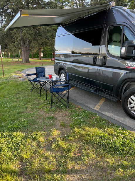Grey RV rental van with extended awning, two blue chairs, and a table set up on the grass.