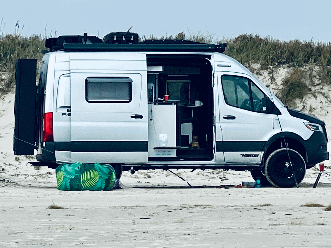 Winnebago RV rental camper van parked on a sandy beach. Its side door is open, showing a cozy interior ready for adventure.