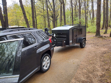 RV rental: dark SUV towing a Hiker Trailer camper & bike. Ready for adventure in a wet, lush forest setting.