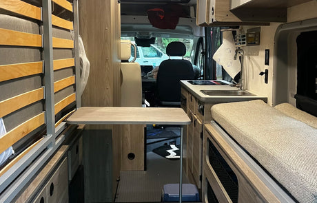 Step inside this rv rental van's compact interior, showcasing a slatted bed, kitchen with sink, and a multi-use table.