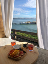 Coastal beauty from an RV rental: a bright ocean view with a pier, framed by white curtains. A wooden table holds a colorful snack board.