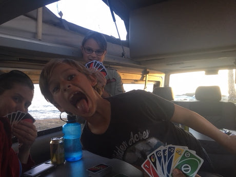 Inside an RV rental, family plays UNO. A boy makes a funny face as kids enjoy cards with an ocean view.