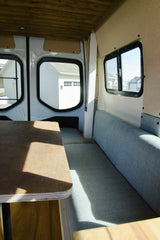 Inside an RV rental: blue upholstered seating, wooden table/ceiling, and windows revealing a residential view outside.