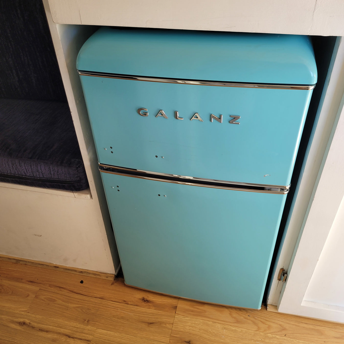 Retro turquoise Galanz mini-fridge built into a cabinet in an RV rental. Features a blue cushion and wooden floor.
