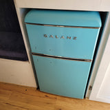 Retro turquoise Galanz mini-fridge built into a cabinet in an RV rental. Features a blue cushion and wooden floor.