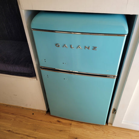 Retro turquoise Galanz mini-fridge built into a cabinet in an RV rental. Features a blue cushion and wooden floor.
