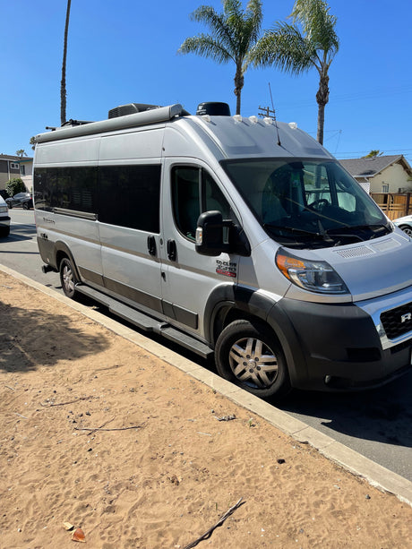 Silver Winnebago Travato rv rental, a Ram ProMaster camper van, parked curbside on a sunny street with sand.