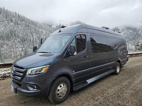 Gray Mercedes-Benz Sprinter RV rental parked amidst snowy mountains and trees on a cloudy day.