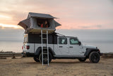 White Jeep Gladiator with rooftop tent. A person looks out over the ocean at sunset, showcasing an ideal RV rental for overlanding adventures.