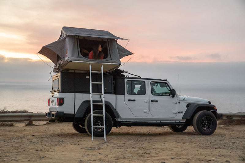 White Jeep Gladiator with rooftop tent. A person looks out over the ocean at sunset, showcasing an ideal RV rental for overlanding adventures. — photo 3
