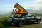 A dark grey Toyota Tacoma with an orange rooftop tent deployed, parked by a misty Pacific coast, showcasing a unique RV rental camping setup.