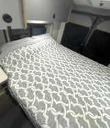 Cozy bed with a grey & white patterned comforter inside an RV rental. Overhead cabinets & window covers visible.