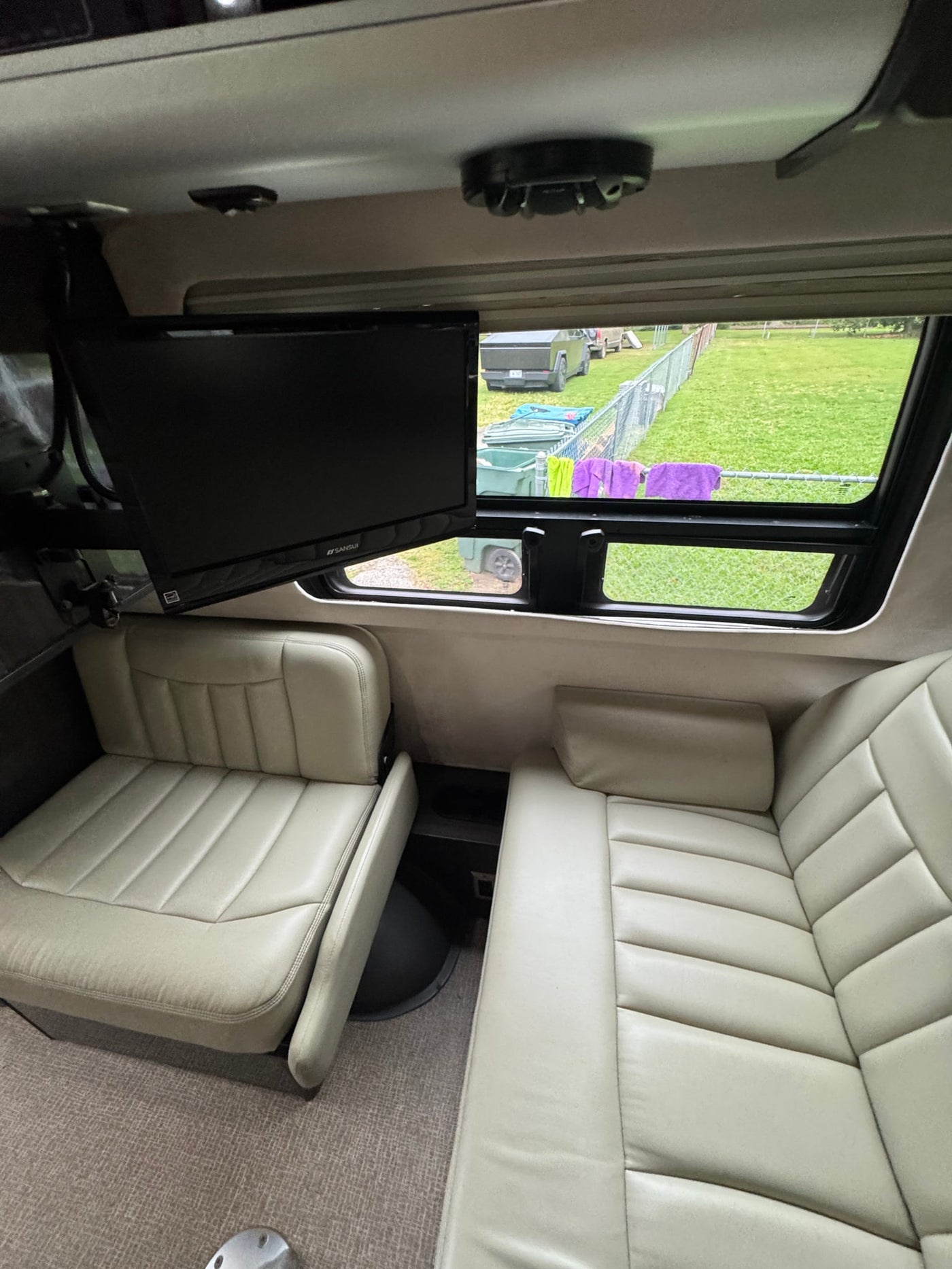 Spacious RV rental interior with light-colored seating, a mounted TV, and a window view of an outdoor field and vehicles. — photo 8