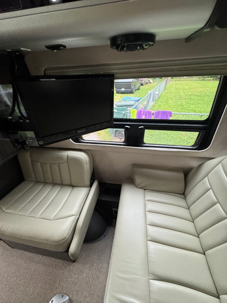 Spacious RV rental interior with light-colored seating, a mounted TV, and a window view of an outdoor field and vehicles.