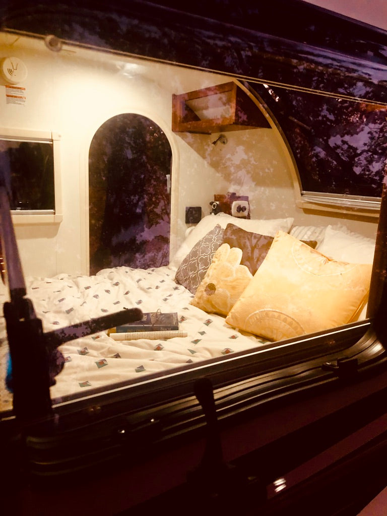 Cozy RV rental interior: A bed with patterned bedding, pillows, and books awaits, featuring an arched doorway and window.