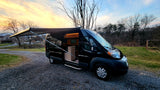A black RV rental van with an extended awning, parked on a gravel drive at sunset. Interior visible, ready for adventures.