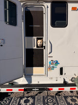 A light-colored dog peeks from the screen door of an RV rental, with scenic travel stickers visible on the camper's side.