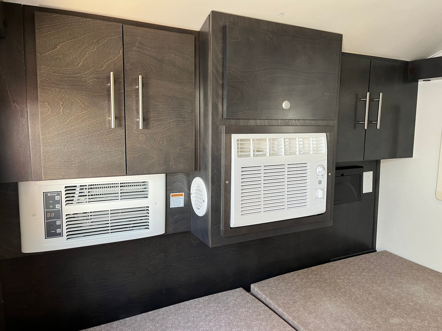 This RV rental's interior features dark wood-grain cabinets housing two white AC/heater units over light cushions, designed for compact comfort.