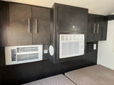 This RV rental's interior features dark wood-grain cabinets housing two white AC/heater units over light cushions, designed for compact comfort.