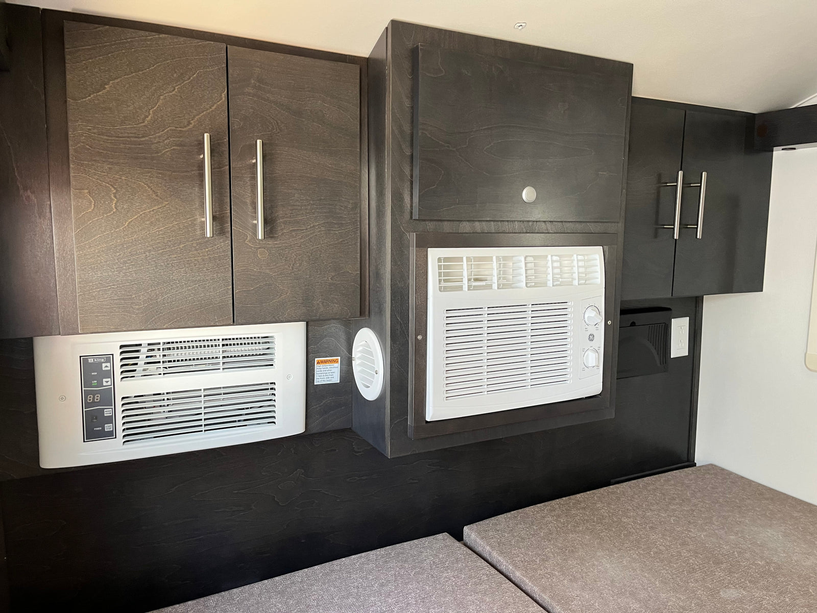 This RV rental's interior features dark wood-grain cabinets housing two white AC/heater units over light cushions, designed for compact comfort.