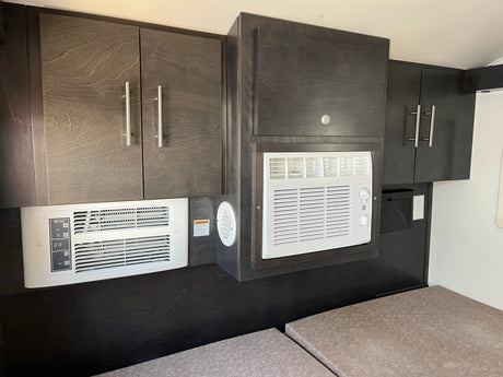 This RV rental's interior features dark wood-grain cabinets housing two white AC/heater units over light cushions, designed for compact comfort.