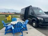 An RV rental, a black camper van, is parked next to blue outdoor chairs and a table at a cloudy campground with a jumping pillow.