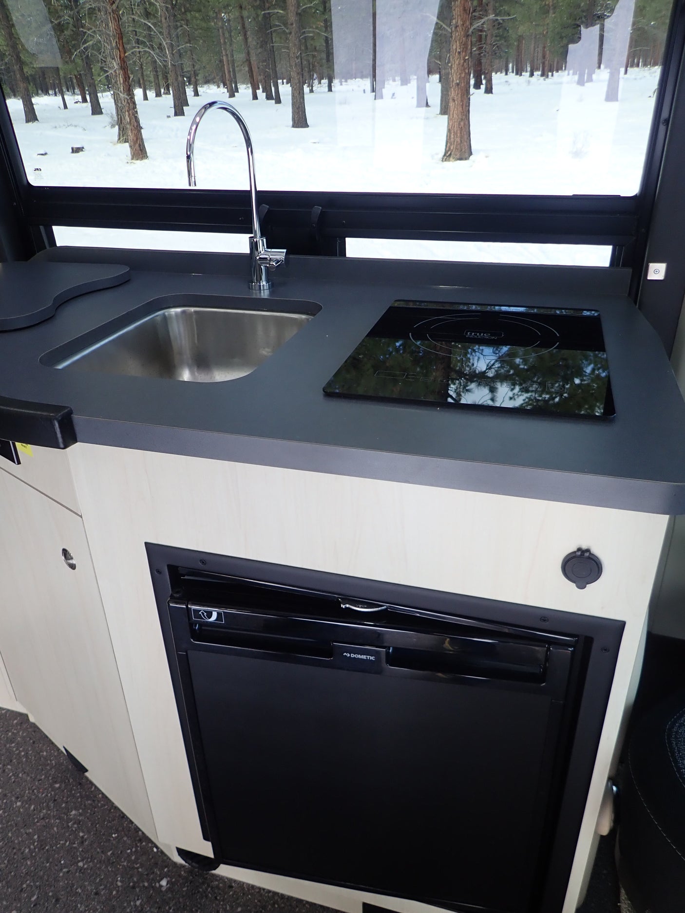 Modern RV rental kitchen features a sink, induction cooktop, Dometic fridge, and a view of a snowy forest.