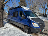 Bright blue Sprinter camper van, a perfect rv rental, parked on a snowy street under a clear winter sky.
