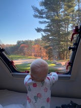 Adorable baby in a fruit-patterned pajama, looking out an RV rental window at vibrant fall trees & a sunny campsite.