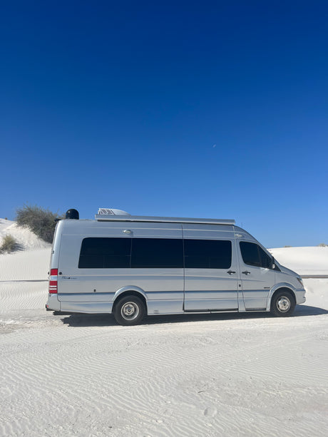 Silver RV rental van parked in vast white sand dunes under a clear blue sky with a crescent moon visible. Perfect desert travel.