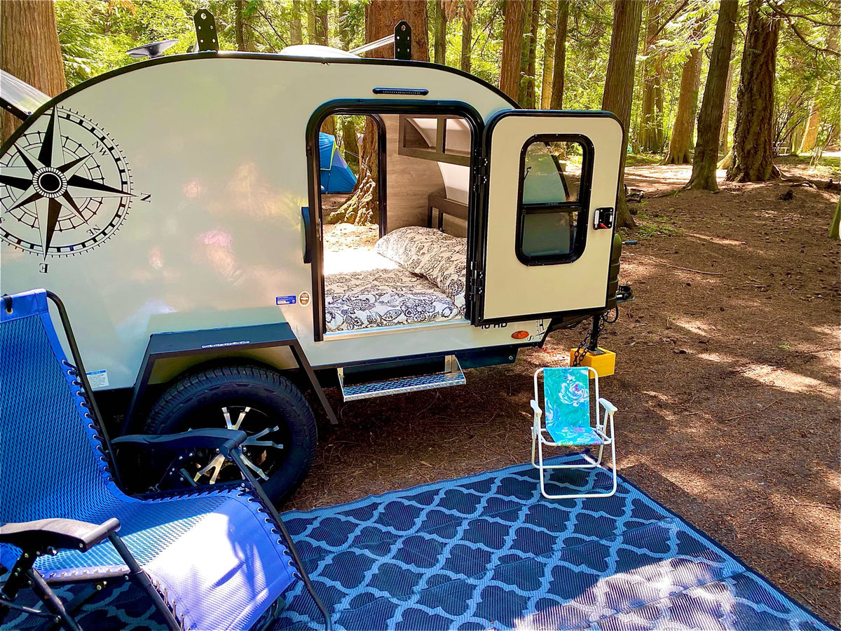 Camping with an RV rental: a compact teardrop trailer, open to a bed, set up with chairs & mat in a sunny forest. — photo 18