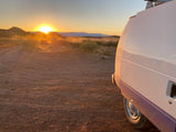Golden desert sunrise viewed from an RV rental, parked on a dusty road amidst scrub brush and distant mountains.
