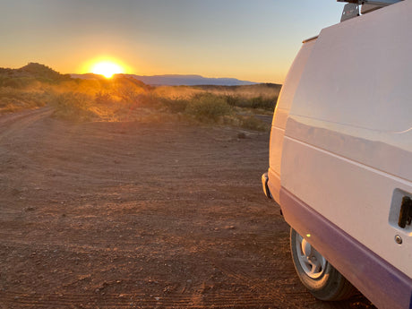 Golden desert sunrise viewed from an RV rental, parked on a dusty road amidst scrub brush and distant mountains.