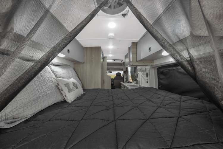 Cozy RV rental interior with a bed, dark comforter, mosquito net, and a glimpse of the kitchen area.
