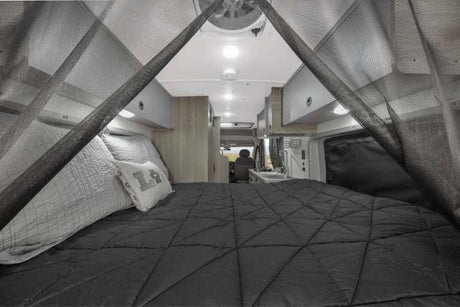 Cozy RV rental interior with a bed, dark comforter, mosquito net, and a glimpse of the kitchen area.