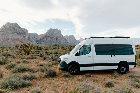 White Sprinter rv rental parked in a vast desert landscape, featuring rugged, layered mountains and desert brush under a cloudy sky.