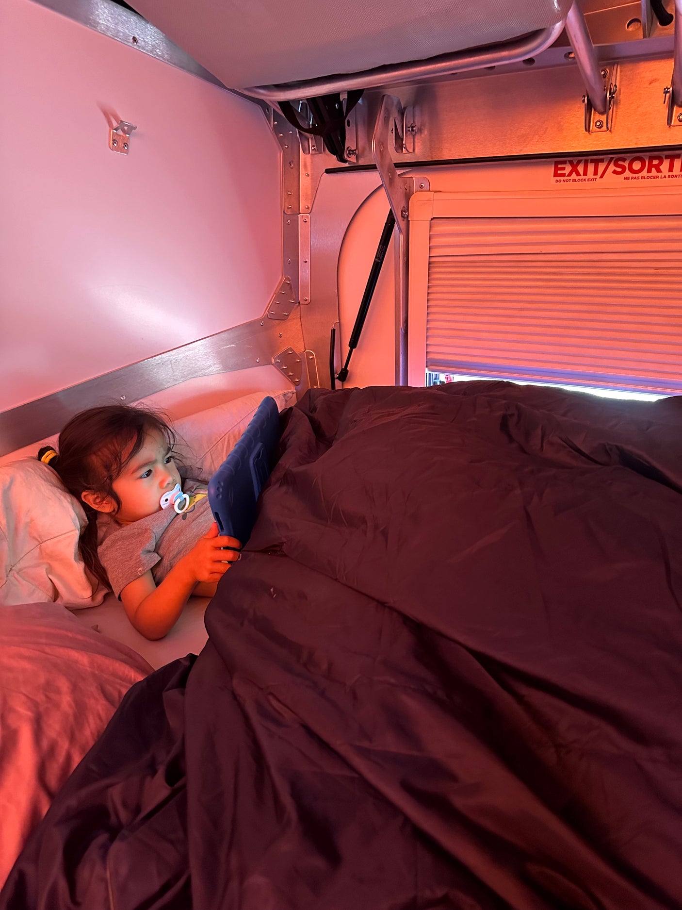Inside an RV rental, a child in bed uses a tablet under warm light, with an "EXIT/SORTIE" sign nearby.