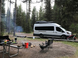 RV rental camper van at a smoky forest campsite. Picnic table cooking, active campfire, and chairs set up for relaxation.